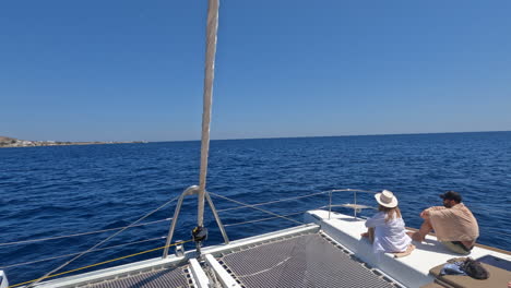 Onboard-a-yacht-sailing-past-the-greek-island-Thera-Santorini-whilst-a-couple-sit-at-the-front-of-the-boat-on-a-beautiful-summers-day