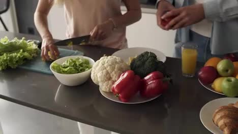 Unrecognizable-young-couple's-hands-cooking-together-a-healty-meal.-Plenty-of-various-colorful-vegetables-on-the-kitchen-counter.-Woman-cutting-salad-on-the-cutting-board,-man-breaking-pepper-slices-on-plate.-Close-up