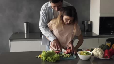 Loving-couple-on-the-grey-kitchen-cooking-together.-Young-woman-shreding-vegetables-together-with-her-husband.-Tall-man-standing-behind-and-teaches-his-wife-to-cut-tomato.-Prepare-lunch-in-romantic-way