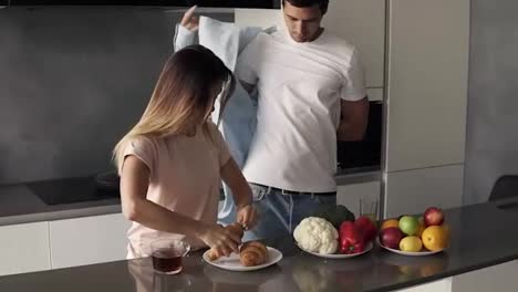 Morning-at-the-kitchen-counter,-fast-breakfast-before-work---croissants-and-coffee-for-breakfast.-Handsome-husband-came-to-the-kitchen-and-putting-on-the-shirt,-in-a-hurry,-wife-eating-croissants