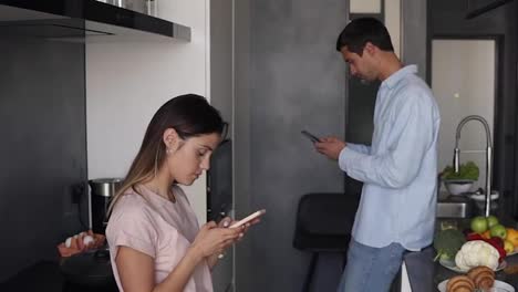 Young-couple-looking-at-something-on-their-smartphones-screen-in-the-kitchen-with-modern-loft-design.-Reading-morning-news-or-checking-social-media