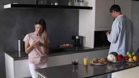 Young-couple-looking-at-something-on-their-smartphones-screen-in-the-kitchen-with-modern-loft-design.-Reading-morning-news-or-checking-social-media.-Standing-separately-from-each-other