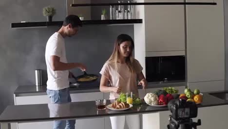 Young-attractive-couple-bloggers-in-casual-clothes-shooting-video-food-blog-about-their-daily-lifestyle,-cooking-simle-breakfast-on-camera-in-the-modern-kitchen