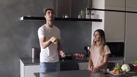 Attractive-loving-caucasian-couple-having-fun-in-the-home-kitchen.-Handsome-man-in-jeans-and-white-T-shirt-juggle-with-apples-to-impress-his-girlfriend,-she-hugs-him.-Slow-motion