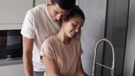 Caucasian-loving-couple-wash-fresh-fruits-in-sink-before-eating-in-the-morning-on-the-modern-kitchen.-Man-in-jeans-gently-stroking-the-back-of-his-caring-wife-then-biting-the-red-apple
