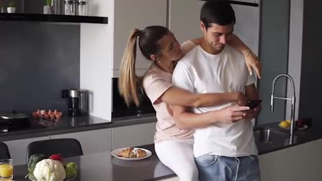 A-man-focused-on-his-phone-while-his-girlfriend-wife-is-trying-to-get-his-attention,-hugs-him-from-the-back.-A-young-couple-standing-embraced-in-casual-clothes-on-the-modern-kitchen