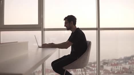 Side-view-of-business-man-using-laptop-computer-at-remote-workplace-in-slow-motion.-Freelancer-working-on-computer-at-home.-Focused-man-looking-laptop-at-big-house-with-panoramic-windows