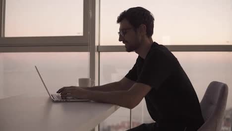 Side-view-of-business-man-using-laptop-computer-at-remote-workplace-in-slow-motion.-Freelancer-working-on-computer-at-home.-Focused-man-looking-on-screen-and-actively-typing-at-big-house-with-panoramic-windows.-Close-up