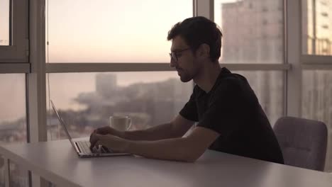 Joyful-man-using-laptop-computer-at-remote-workplace-in-slow-motion.-Freelancer-working-on-computer-at-home.-Good-looking-man-actively-typing-on-laptop,-chatting-with-friends-at-big-house-with-panoramic-windows.-Close-up