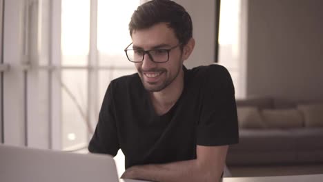 Portrait-of-a-toothy-smiling-man-in-glasses-using-laptop.-Freelancer-working-on-computer-at-home.-Handsome-man-actively-typing-on-laptop,-chatting-with-friends-at-big-house-with-panoramic-windows-and-sofa-on-background