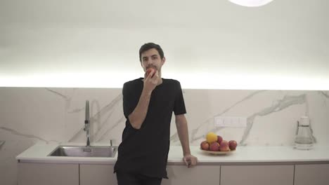 Young-man-eating-red-apple-at-home-in-the-kitchen.-Portrait-of-guy-in-black-clothes-eating-a-fresh-apple-and-looking-at-camera.-Leaning-on-a-counter-at-modern,-white-kitchen