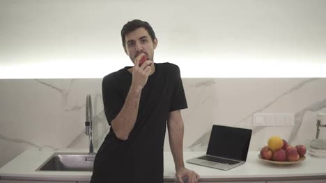 Young-man-eating-red-apple-at-home-in-the-kitchen.-Portrait-of-guy-in-black-clothes-eating-a-fresh-apple-and-looking-at-camera,-smiling.-Leaning-on-a-counter-at-modern,-white-kitchen,-laptop-next-to-him