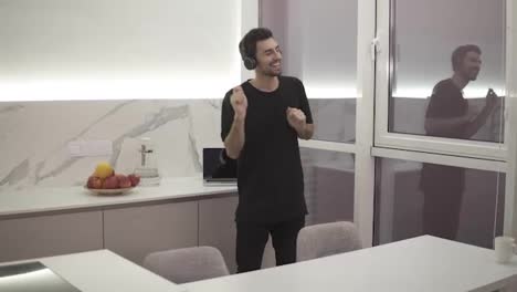Joyful-man-in-black-clothes-is-dancing-in-kitchen-listening-to-music-with-headphones,-having-fun-alone-in-light-modern-apartment.-Devices-and-people-concept.-Accelerated