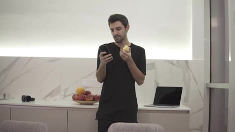 Handsome-man-browsing-on-mobile-phone-at-home-bright-kitchen.-Handsome-young-man-browsing-on-smartphone-smiling-happy.-A-man-eats-an-apple