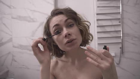 Close-up-shot.-Beautiful-young-woman-putting-mascara-and-looking-at-camera.-White-marble-interior-in-bath-room.-Beauty-and-makeup-concept.-Slow-motion