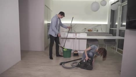 Cleaning-day.-Young-couple-preparing-tools-for-cleaning-their-apartment---man-taking-wiping-mop,-girl-preparing-vacuum-cleaner.-Gonna-clean-studio-kitchen-together