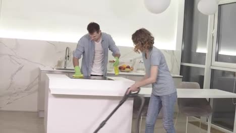 Cleaning-day.-Young-couple-cleaning-their-apartment---man-in-gloves-wiping-up-kitchen-stove,-girl-cleaning-floor-using-vacuum-cleaner.-Clean-studio-kitchen-together-in-the-evening.-Smiling-to-the-camera