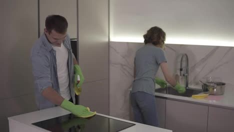 Young-smiling-married-couple-in-casual-clothing-and-both-in-green-gloves-cleaning-kitchen-after-moving-to-new-home,-household.-Woman-is-washing-the-dishes,-man-is-cleaning-a-kitchen-stove-with-a-mop-and-chemical-spray