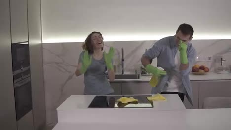 Happy-young-couple-dancing-in-kitchen-both-in-green-rubber-gloves-having-fun-on-clean-up-day-in-studio-apartment.-Modern-youth,-people-and-housekeeping-concept