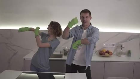 Happy-young-couple-dancing-synchronously-in-kitchen-both-in-green-rubber-gloves-having-fun-on-clean-up-day-in-studio-apartment.-Funny-moves.-Modern-youth,-people-and-housekeeping-concept