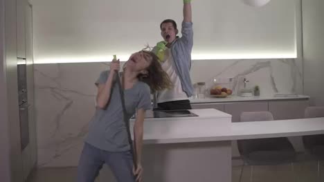 Two-young-people-dancing-together-and-emotionally-singing-using-broom-and-cleaning-tools-as-as-microphones-while-cleaning-in-kitchen.-Happy-couple-enjoying-time.-Slow-motion