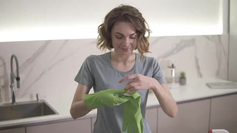 Portrait-of-determined-household-ready-to-start-cleaning-the-household,-puts-on-green-gloves-and-smiling-and-looking-to-the-camera.-Bright,-modern-kitchen.-Slow-motion
