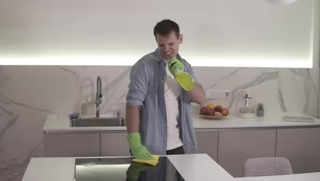 Tall-man-in-green-protective-gloves-is-singing-using-a-chemical-spray-as-microphone-while-cleaning-the-ceramic-stove-surface-using-mop.-Emotional-singing-during-cleaning