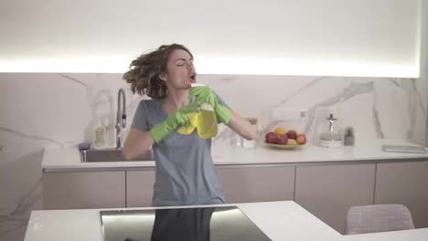 Cute-short-haired-woman-in-green-protective-gloves-is-singing-using-a-chemical-spray-as-microphone-while-cleaning-the-ceramic-stove-surface-using-mop.-Emotional-singing-during-cleaning