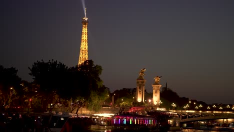 Eiffel-Tower-seen-at-night-behind-the-Alexandre-III-Bridge-in-the-Seine-river-with-party-boat-below,-View-from-tour-boat