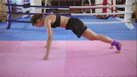 Athletic-woman-in-bra-and-shorts-standing-on-her-knee-and-fists-then-starting-to-do-push-ups-on-exercise-mat-inside-of-the-boxing-ring.-Tough-power-training.-Shot-in-4k