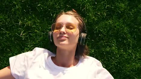 Girl-listening-to-music-streaming-with-headphones-in-summer-on-a-meadow.-Green-grass.-Smiling-girl-in-yellow-sunglasses-and