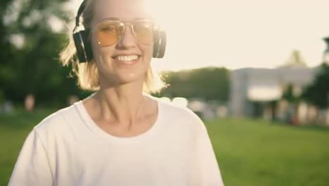 Attractive-caucasian-girl-having-fun-outdoors.-Posing-and-smiling-to-camera-wearing-white-T-shirt,-headphones-on-her-neck