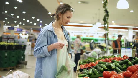 Shopping-in-the-supermarket.-Young-woman-in-casual-in-vegetables-department.-Selecting-fresh-cucumbers.-Puts-them-into-the-cellophane-bag.-Blurred-background