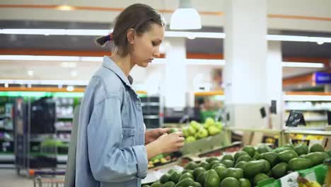 Vegetable-and-fruits-department-in-the-grocery-store.-Pretty-girl-in-casual-selecting-green-avocado.-Handheld-footage.-Side-view