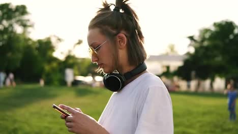 Side-view-of-a-fancy-young-girl-in-white-T-shirt-typing-on-her-mobile-phone.-Black-headphones-on-her-neck.-Spending-time-in-the