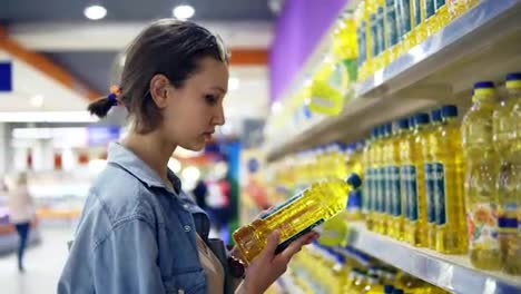 Girl-in-the-store,-reading-information-sticker-on-an-oil-bottle.-Selected-one-bottle-of-sunflower-oil,-puts-it-n-trolley.-Assortment-of-products-in-the-row