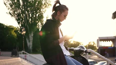 Young-girl-in-black-hoodie-sitting-on-a-motorbike-and-typing-on-her-mobile.-Sitting-on-moped-outside-in-the-park.-son-rays-on