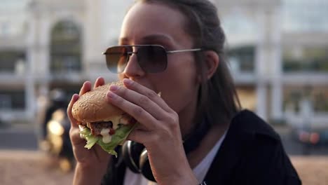 Attractive-young-woman,-smiling-cheerfully,-holds-tasty-burger-in-two-hands-and-eats-it.-Dressed-in-casual-outfit,-in-sunglasses
