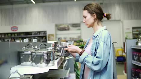 A-woman-in-a-store-in-casual-clothes.-Standing-near-a-shelf-with-pots,-examines-products.-Selecting.-Kitchen-stuff.-Housewife-in-supermarket.-Side-view