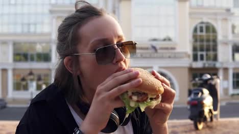 Young-female-eats-a-big,-juicy-hamburger-with-two-hands.-Young-girl-with-short-hair-and-dark-sunglasses.-Hungry.-Close-up