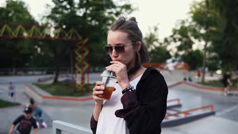 A-stylish-girl-in-sunglasses-and-headphones-enjoys-a-drink-in-a-transparent-glass,-drinks-through-a-straw.-A-woman-stands-in-the