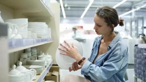 A-girl-in-a-shop-looks-at-white-ceramic-dishes.-She-turns-the-plate-in-her-hands,-chooses.-Looks-at-the-dishes-from-the-bottom-shelf-in-the-store.-Came-down.-Assortment-of-products
