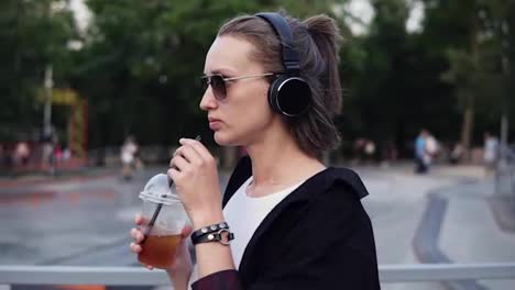 Side-view-of-caucasian-girl-walking-through-park-side-and-drinking-a-soft-drink-with-a-straw.-Walk-in-sunglasses,-black-hoodie