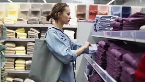 Wide-assortment-of-different-colors-bath-towels.-Side-view-of-a-female-buyer-between-the-rows.-Unfolds-the-towels.-Selecting.-Supermarket