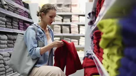 Caucasian-woman-choosing-towels-from-big-variety-in-the-row.-Shelves-in-store-are-fulled-of-different-towels-of-any-color.-Girl-unfolds-one-big-red-towel-from-the-bottom-shelf