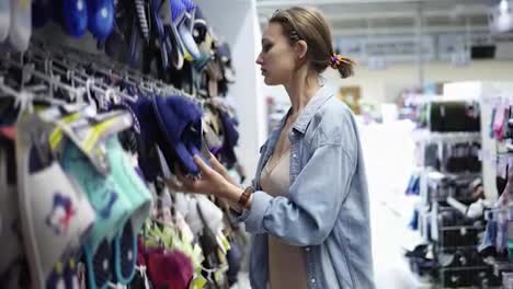 Pretty,-caucasian-girl-in-a-store-between-rows-of-different-stuff.-Choosing-slippers-from-a-big-assortment.-Girl-in-a-casual-clothes,-housewife-shooping.-Side-view