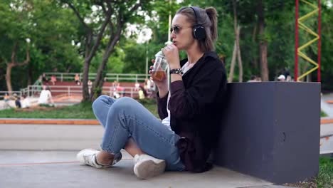 Stylish,-young-girl-sits-in-a-park-on-the-floor-with-her-legs-crossed.-Sipping-a-soft-drink.-A-woman-with-sunglasses-and
