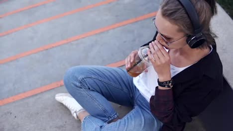 High-angle-footage-of-a-beautiful-girl-sitting-on-the-ground-or-asphalt-drinking-cocktail-from-the-transparent-glass-using-a