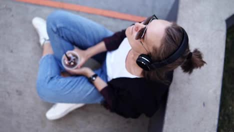 Relaxed-young-woman-sitting-on-the-floor-outdoors-and-listening-to-the-music.-Wearing-blue-jeans,-white-T-shirt-and-black-coat