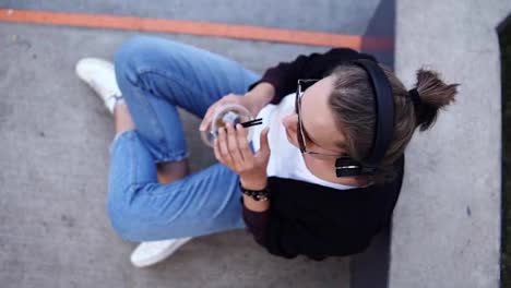Attractive-girl-in-sunglasses-and-headphones-sitting-on-the-concrete-surface-with-crossed-legs.-Footage-from-the-top.-Girl